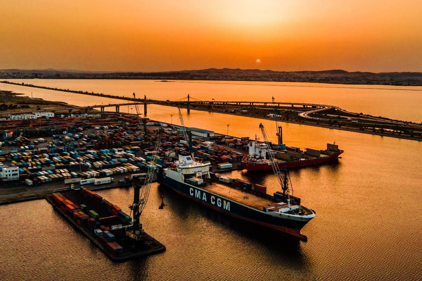 A container vessel berthed at Radès port, Tunis, at dusk, with the container yard alongside and the road bridge crossing the canal beyond.